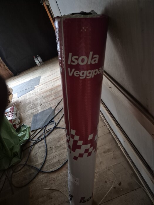 Rulle med Isola väggpapp stående i ett rum under renovering, omgiven av kablar och byggmaterial på trägolv.