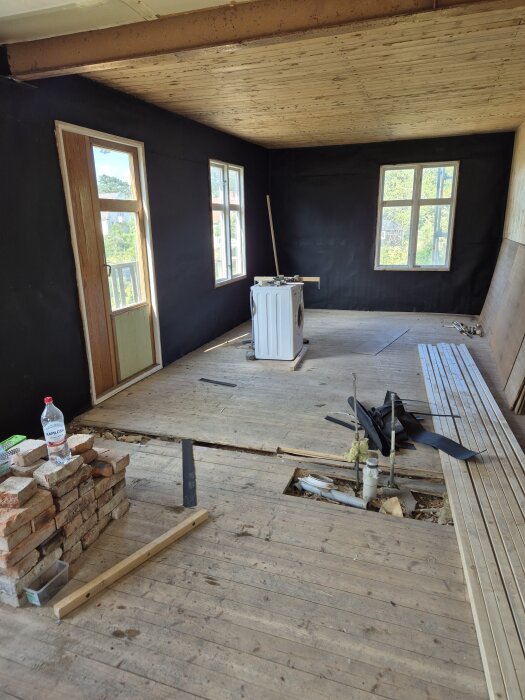 Renoverat rum med svart väggpapp, blottat tak och golv, en stapel tegelstenar, och verktyg utspridda. Förberedelse för ytterligare arbete.