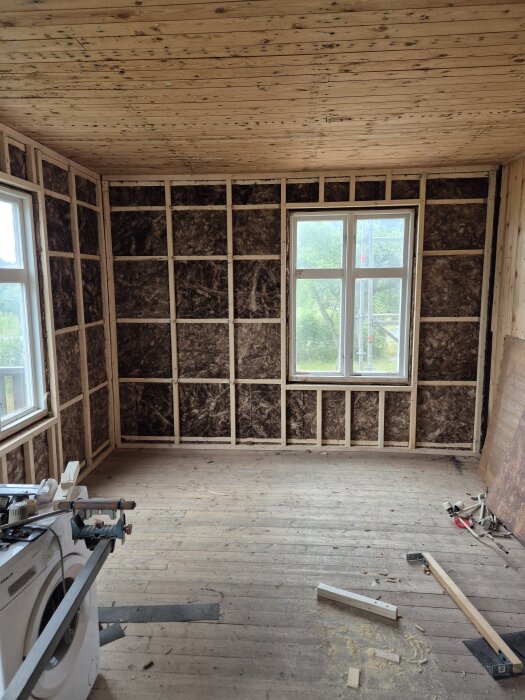 Ett rum under renovering med träreglar och isolering i väggarna, två fönster släpper in ljus. Golvet är delvis täckt med verktyg och material.