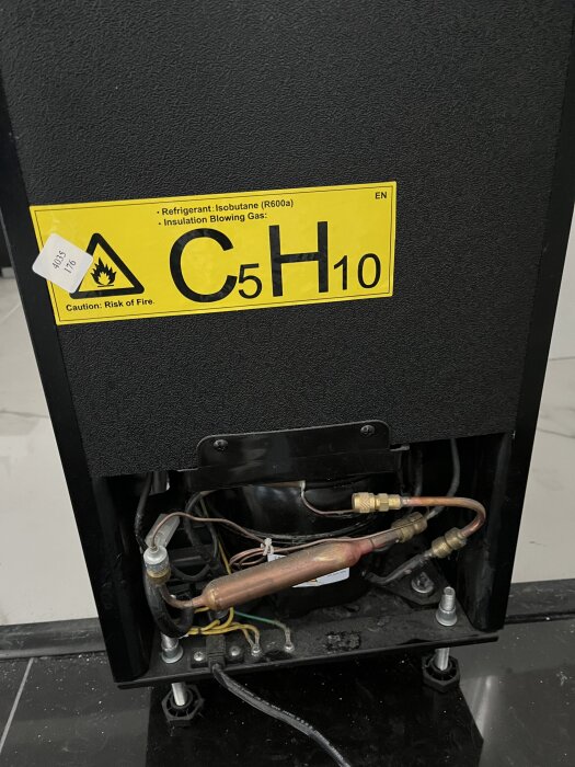Baksidan av en kompressor, synliga rör och etikett med text om isobutan, säkerhetsvarning och risken för brand, i samband med kylsystemfråga.