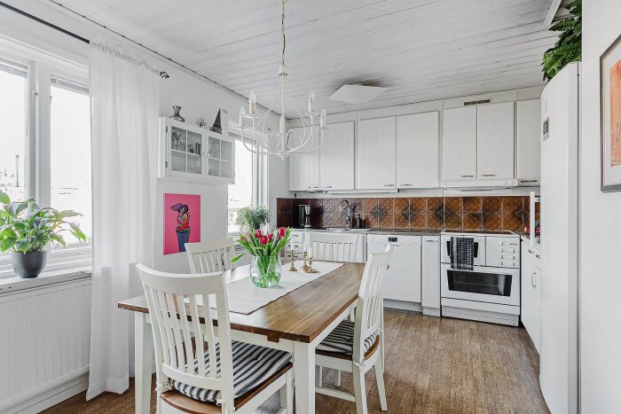 Ljust kök med vita skåp, träbord med blommor, och konstverk på vägg. Rummet har mycket naturligt ljus från stora fönster.