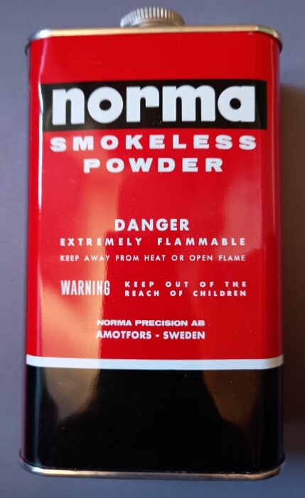 Röd och svart behållare märkt "Norma Smokeless Powder" med varningstexter om brandfara och barnsäkerhet.