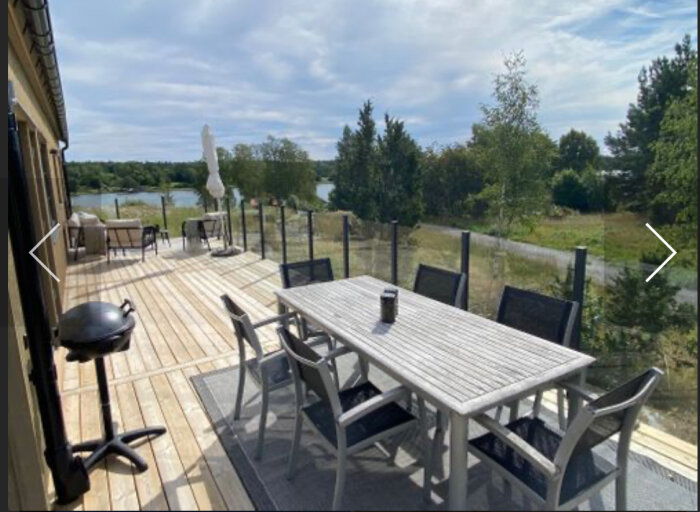 Stor träaltan med utemöbler, grill och glasräcke, omgiven av grönska och sjöutsikt.