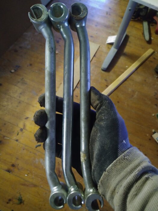 Tre metallverktyg hålls i en handskbeklädd hand på ett trägolv.
