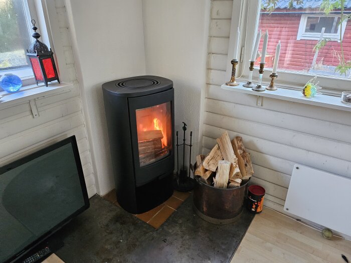 Svart braskamin Contura 550 med eld, vedkorg och värmeplatta, placerad i ett hörn med vita väggar och en fönsterkarm med ljusstakar.