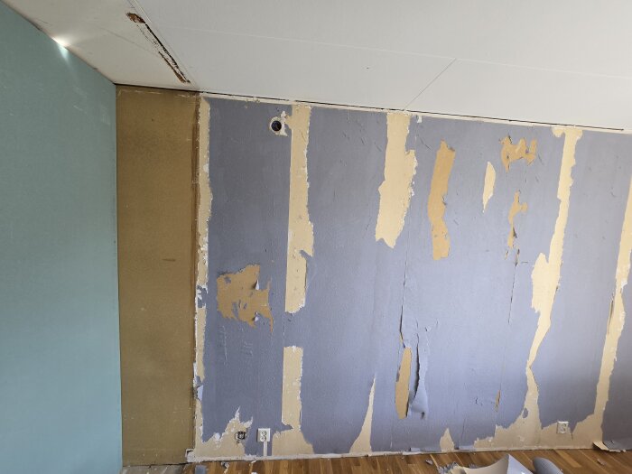 Vägg i rum under renovering, avskavd tapet på höger sida och turkos vägg till vänster. Golvet är trä och eluttag syns längst ner.