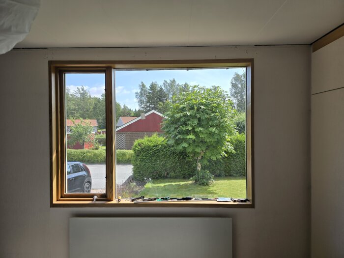 Vy från ett fönster mot en trädgård med ett träd, buskar och en bil parkerad på uppfarten, omgiven av röda hus och skog i bakgrunden.