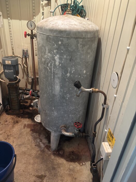 Gammal hydrofor med rörsystem, kontrollventiler och tryckmätare. Bottenventil för tömning och rengöring visas.