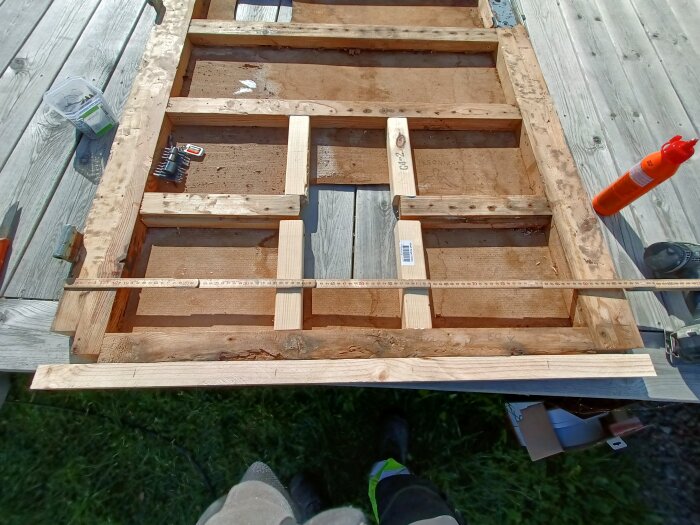 Träram med tvärslåar i byggprocess, måttstock på ovansidan och verktyg runtom, liggande på träbord utomhus.