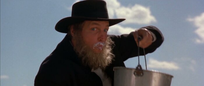 Gammaldags klÀdd man med skÀgg och mjölk kring munnen hÄller en hink, under blÄ himmel.