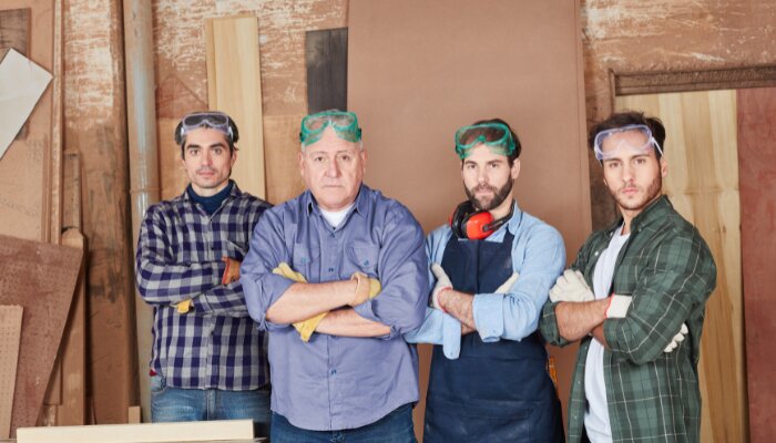Fyra hantverkare i arbetskläder och skyddsglasögon står med armarna i kors i ett snickeri.