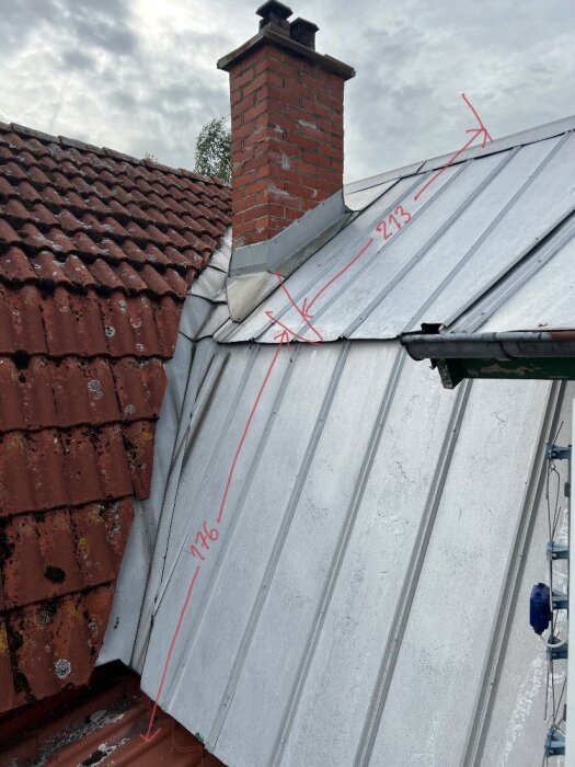 Plåttak med veckade långsidor bredvid ett gammalt tegeltak, röd tegelstensmur, och handskrivna måttangivelser i röd färg på plåttaket.