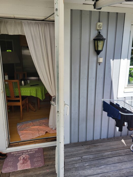 Öppen altandörr in i ett rum med gardiner och grön bordsduk, utanför en trätrall och en lampa på väggen, kläder hänger på en torkställning.