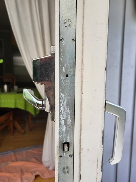 Närbild av lås och handtag på en vit altandörr med metallbeslag. Ett rum med grönt bord syns i bakgrunden.