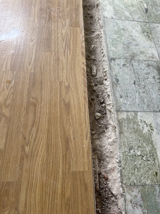 Trägolv intill ett golv av Kolmårdenmarmor med synlig nivåskillnad och ojämn kant. Mark mellan golven visas.
