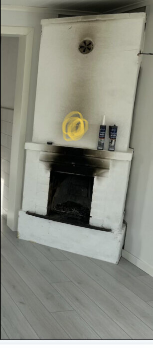 Vit murad öppen spis med sotfläckar upptill och några tuber tätningsmassa ovanpå spishyllan. Golvet är av grå trälaminat.