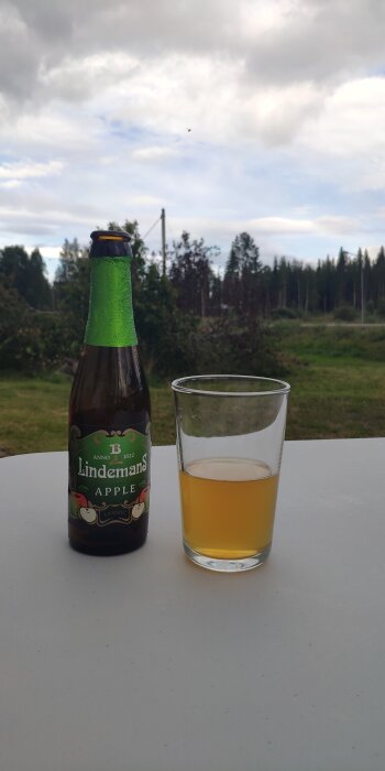 Flaska med Lindemans äppelöl och ett glas med öl på ett bord utomhus, grönskande landskap i bakgrunden.