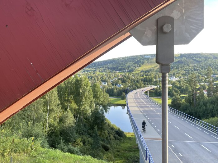 Röd brobjälke i förgrunden, med väg och cyklist på bro över frodig dal och flod. Skog och ås i bakgrunden under klar himmel.