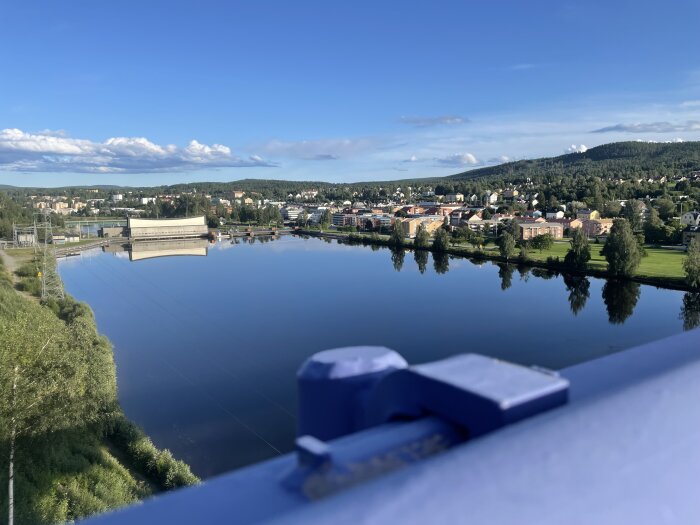 Utsikt från en bro över en stilla flod med spegelblanka vattenytor, omgivande grönska och en stad i bakgrunden under en klarblå himmel.