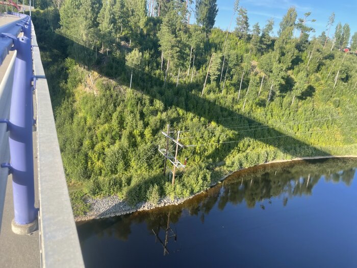 Bro över en flod med grön skog i bakgrunden. Elstolpar med ledningar korsar landskapet. Solig dag med blå himmel.