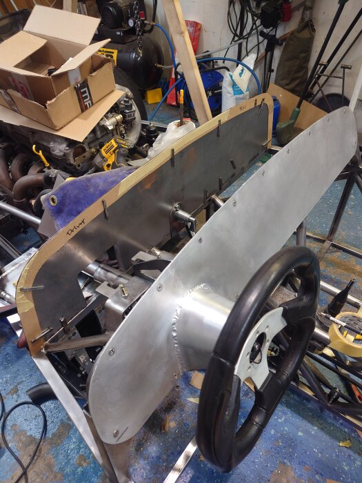 Bilens instrumentpanel under konstruktion med flera fästhål. En ratt och verktyg synliga i bakgrunden. Projektmaterial och kartonger runt omkring.