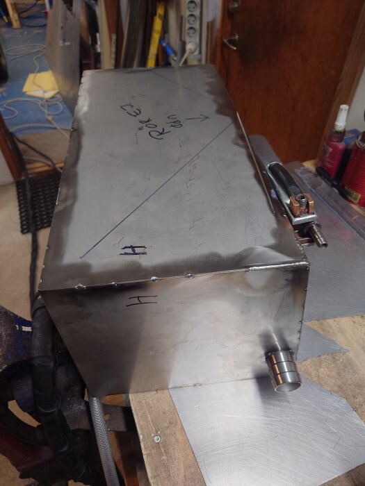 Metallåda under konstruktion med handskrivna markeringar och svetsade kanter, omgiven av verkstadstillbehör.