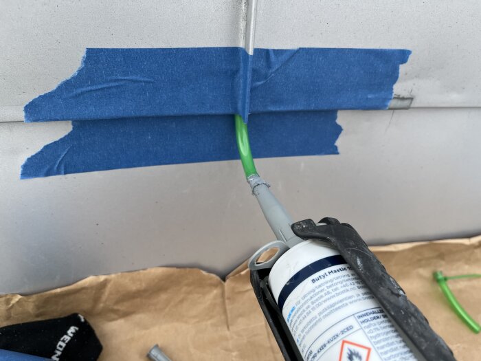 Användning av butylmassa vid rörinstallation, där en grön slang fästs på en metallplåt med blå tejp och butyl injiceras med en spruta.