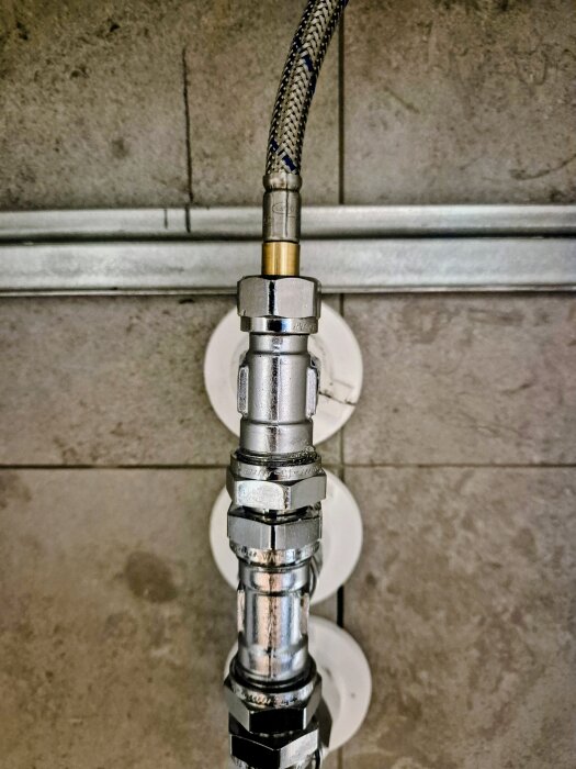 Närbild av vattenslanganslutning med metallkopplingar och ventiler, monterad på en kaklad vägg.