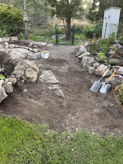Trädgårdsgång med jord och stenar kantad av växter, leder till en grön grind. Skyfflar står på marken, omgivande skog i bakgrunden.