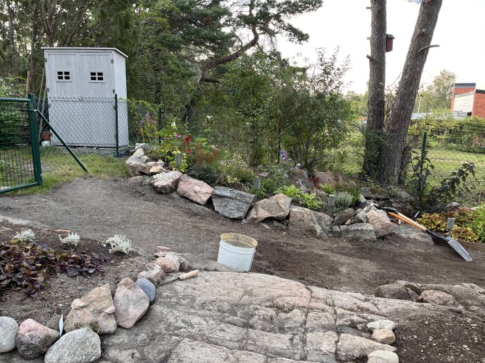 En grusgång i trädgården kantad av stenar, omgiven av växter och träd, med en grind och ett litet skjul i bakgrunden.