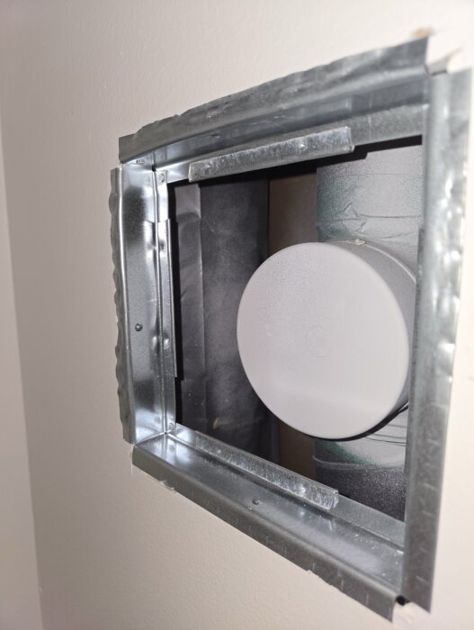 Inspektionslucka i vägg med synlig frånluftsrör, omgiven av isolering. Ramen är av metall och har en plastig utseende runt röret.