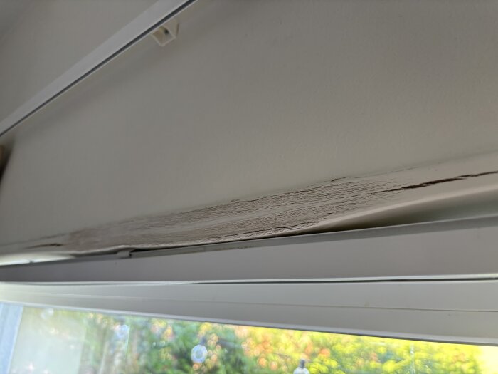 Svullet fönsterfoder ovanför fönster orsakat av vatteninträngning efter kraftigt regn, utan andra synliga skador på väggen eller fasaden.
