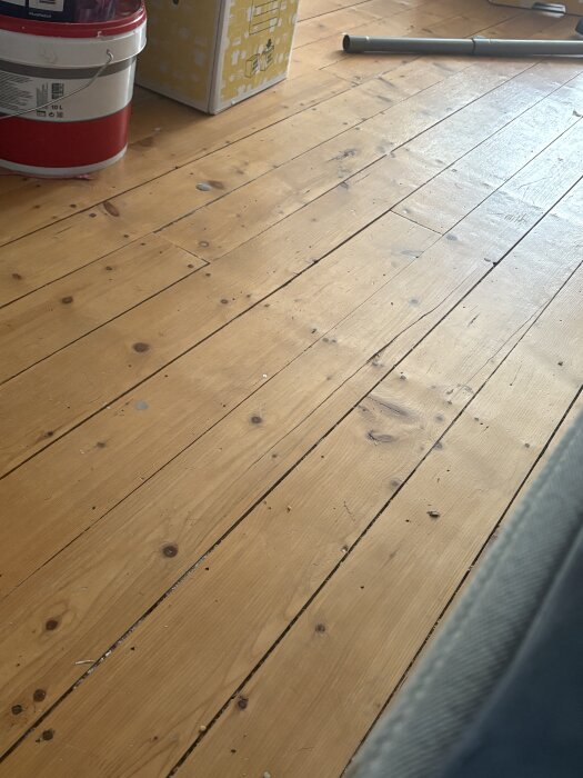 En nyslipad och lackerad trägolv med soliga reflektioner syns, omgiven av en färgburk, en kartong och en rördel.