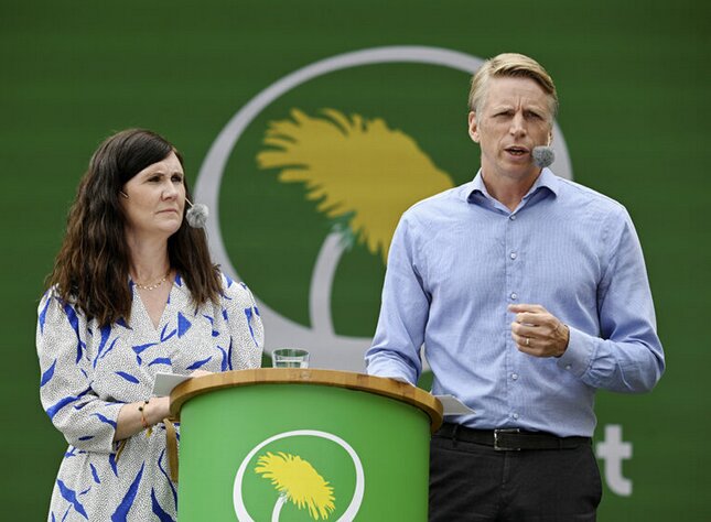 Två personer står vid ett podium med en grön bakgrund och en mikrofon, under en presentation eller tillkännagivande.