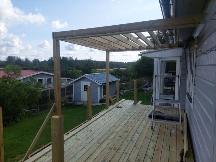 Träpergola under konstruktion på en terrass bredvid ett hus, med grönska och små byggnader i bakgrunden.
