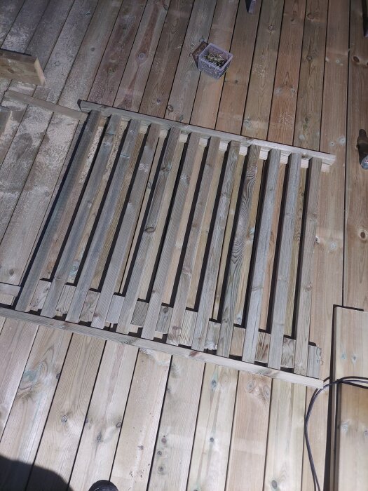 Träribbor ligger på ett bygggolv, del av pågående arbete med en pergola eller staket. En låda med skruvar syns i bakgrunden.