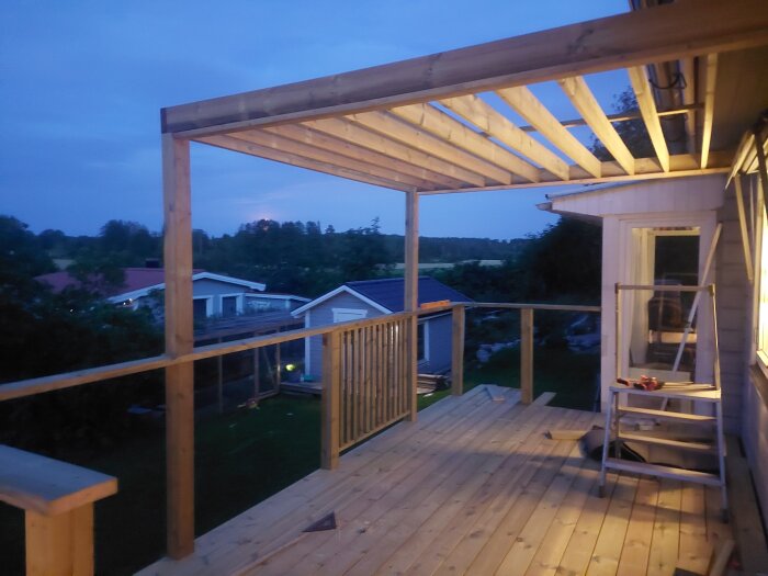 Pergola med takbjälkar och räcke byggs på en altan i skymning, med utsikt över trädgård och hus. Verkyg och stege syns på altangolvet.