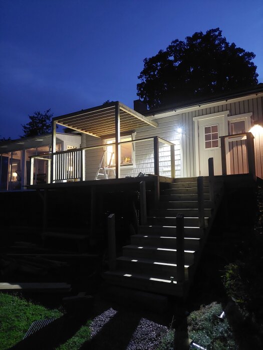 Pergola under konstruktion vid ett hus på kvällen, med överliggare och sektioner för räcket synliga i ljuset från inomhusbelysning.