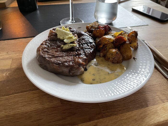 Entrecote med örtsmör, ugnsrostad potatis och tomatskivor samt en krämig sås på en tallrik.