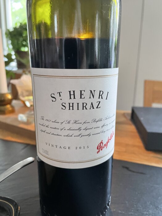 Flaska med St. Henri Shiraz rödvin, årgång 2015, från Penfolds. Fotot togs inomhus på ett bord med suddig bakgrund.