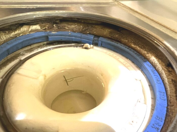 Golvbrunn med synlig blå brunnsmanchett och brun beläggning runt kanterna i ett badrum.