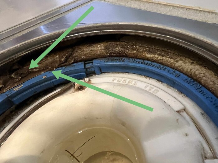 Närbild av en golvbrunn med gröna pilar som pekar på brun kant, misstänkt fogmassa eller lös brunnsmanschett, delvis synlig blå ring.