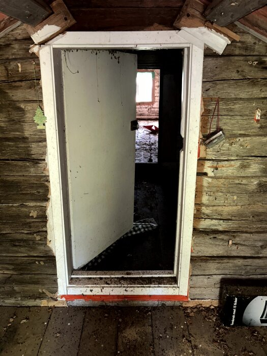 En dörröppning i en gammal timmerstuga med en vit dörr som står på glänt, synlig innervägg av timmer och en öppning mot rummet bakom.