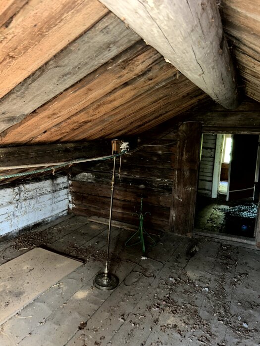 Gammal torpinredning, lågt tak med träbalkar, slitet trägolv, lampa och några föremål i hörnet, antydan till öppning mot annat rum.