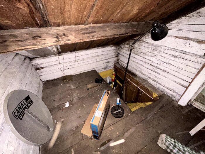 Ett trångt loft med synliga takbjälkar, trägolv, en golvlampa, en Canal Digital-parabol och diverse föremål som en pappkartong och brandsläckare.