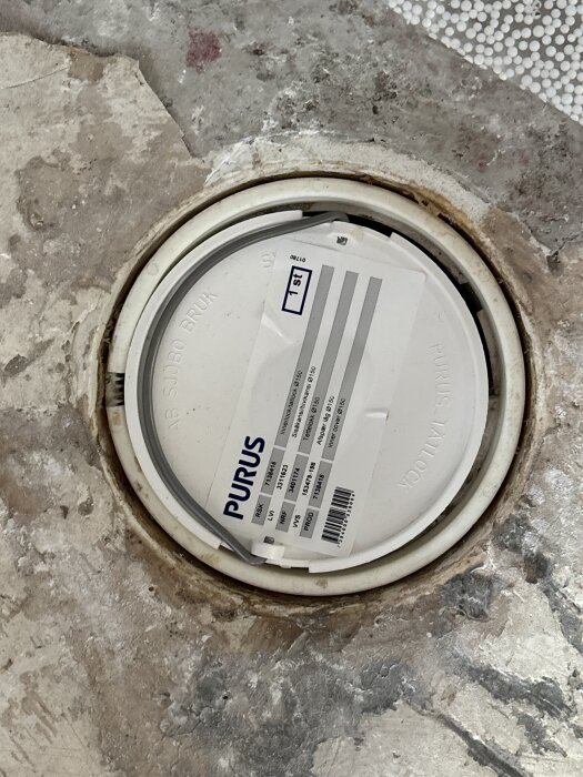 Golvbrunn med vitt tätningslock från Purus på betonggolv, med text och streckkod synliga på locket.