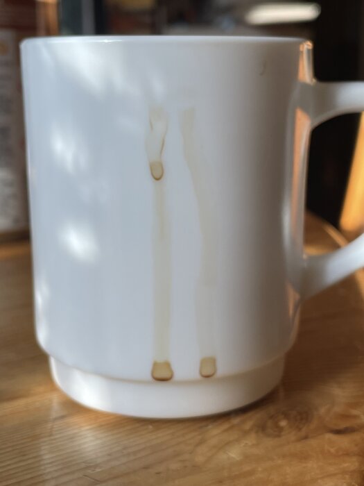 En vit kaffekopp med kaffefläckar på utsidan, stående på ett träbord i naturligt ljus.
