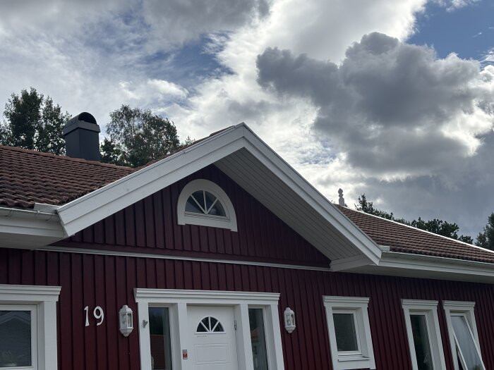 Rött hus med vit dörr och fönster, sadeltak och en skorsten. Molnig himmel i bakgrunden.