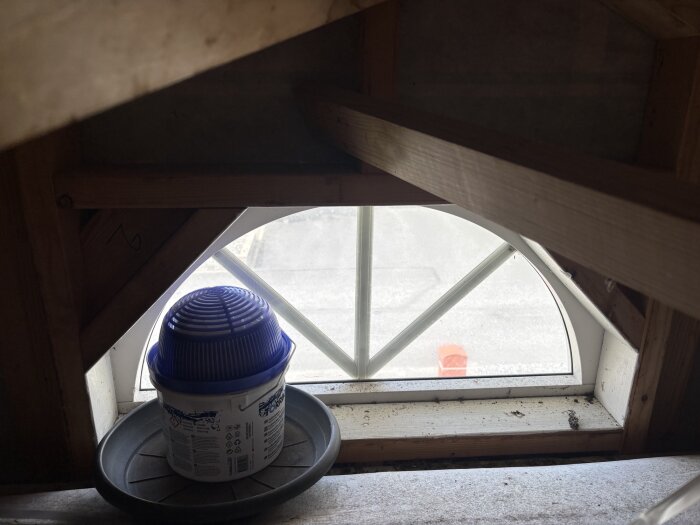 Plastråg med avfuktare står framför ett litet fönster i en utbyggnad på vinden, omgivet av träbalkar.