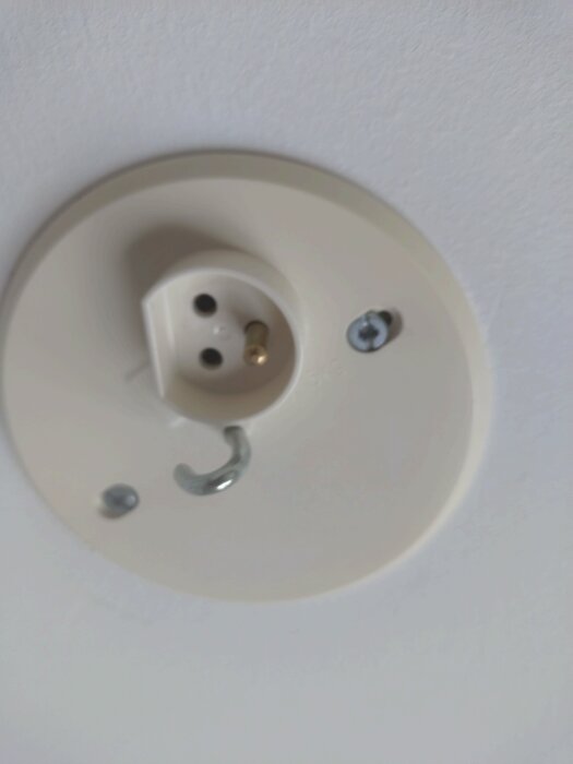Vit lamphållare i tak med metallkrok och skruvar, utan kåpa. Problem med att stabilt fästa lampkåpan nämns i diskussionen.